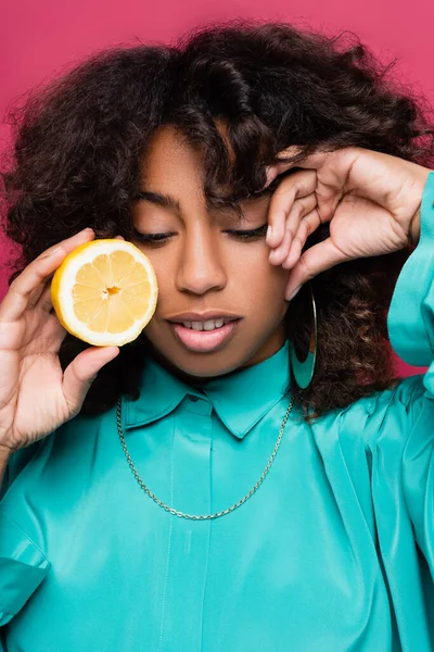 Elegante mujer afroamericana con limón fresco y mano cerca de la cara aislado en rosa - foto de stock