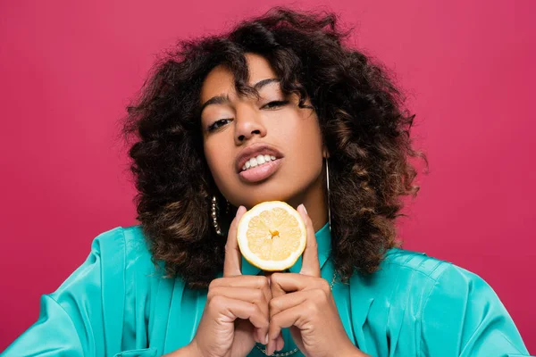 Mujer afroamericana rizada sosteniendo medio limón y mirando a la cámara aislada en rosa - foto de stock