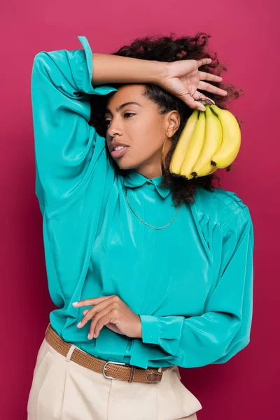 Mujer afroamericana en blusa turquesa posando con rama de plátano aislada en rosa - foto de stock