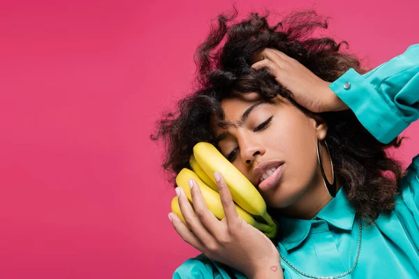 African american woman touching curly hair while posing with bananas near face isolated on pink — Stock Photo