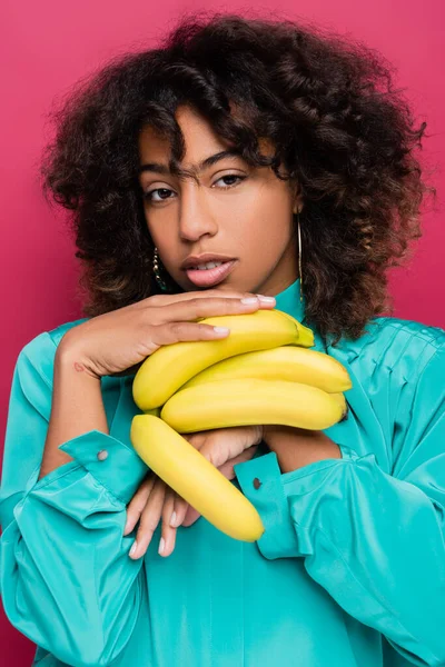 Mujer afroamericana rizada con plátanos maduros mirando a la cámara aislada en rosa - foto de stock