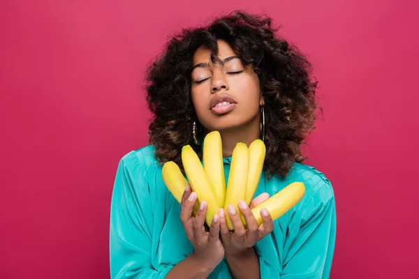 Mujer afroamericana de moda con los ojos cerrados posando con plátanos aislados en rosa - foto de stock