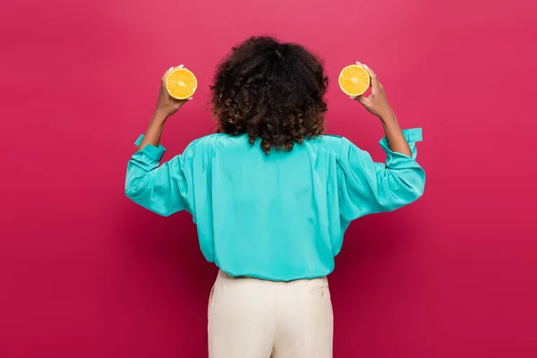 Vista posterior de la mujer afroamericana en blusa azul sosteniendo mitades anaranjadas aisladas en rosa - foto de stock