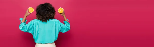 Back view of curly african american woman with halves of ripe orange isolated on pink, banner — Stock Photo