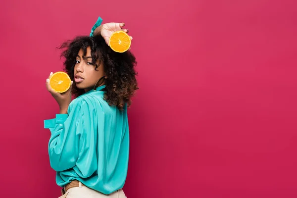 Mulher americana africana encaracolado com metades de laranja suculento olhando para a câmera isolada em rosa — Fotografia de Stock