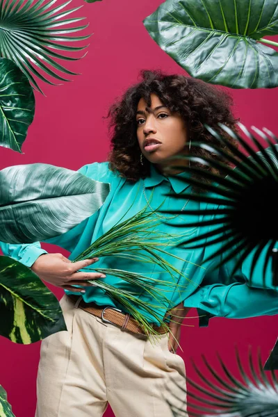 Donna afroamericana alla moda in posa con mano in vita vicino alle foglie tropicali isolate sul rosa — Foto stock