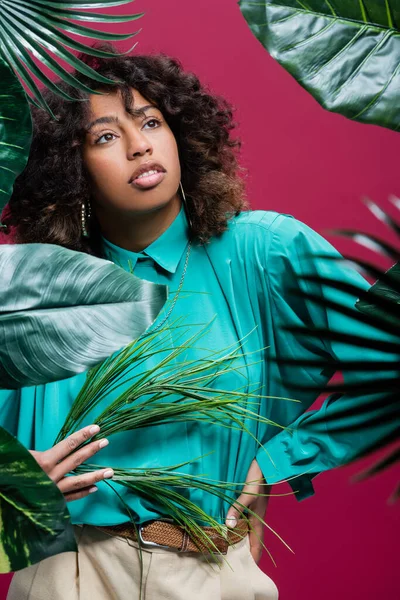African american woman in turquoise blouse standing with hand on waist near exotic leaves isolated on pink — Stock Photo