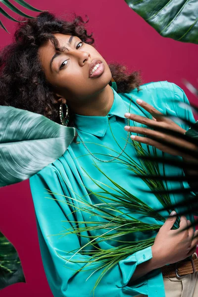 African american woman in stylish turquoise blouse posing near exotic leaves isolated on pink — Stock Photo