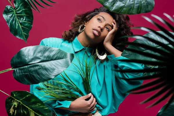 Femme afro-américaine en chemisier turquoise tenant la main près du visage tout en posant avec des feuilles tropicales isolées sur rose — Photo de stock