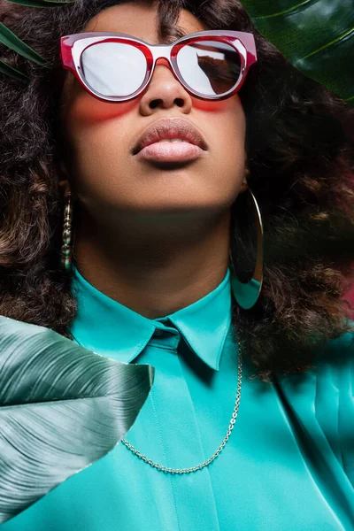 Femme américaine africaine bouclée dans des boucles d'oreilles et des lunettes de soleil près de feuilles vertes — Photo de stock