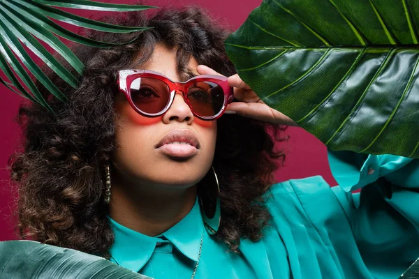 Afro-américaine femme ajuster lunettes de soleil à la mode près de feuilles exotiques isolées sur rose — Photo de stock