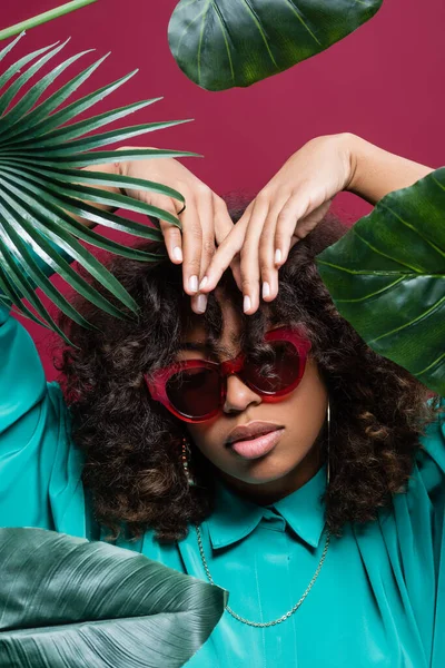 African american woman in sunglasses posing with hands above head near leaves isolated on pink — Stock Photo