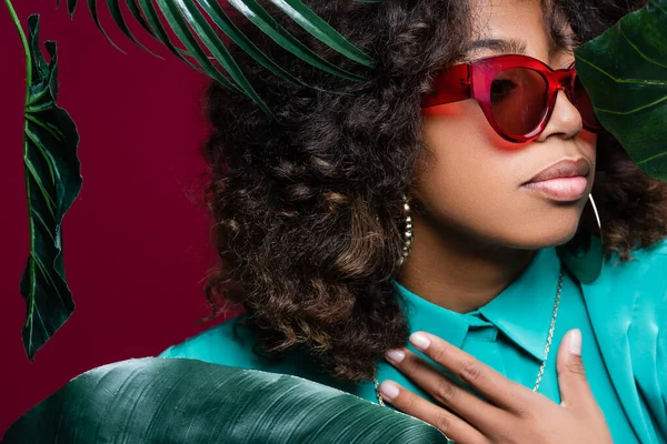 Brunette afro-américaine en lunettes de soleil touchant la poitrine près de feuilles vertes isolées sur bordeaux — Photo de stock