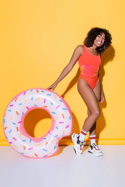 Pleine longueur de femme afro-américaine en maillots de bain et baskets près de l'anneau de natation sur fond jaune — Photo de stock