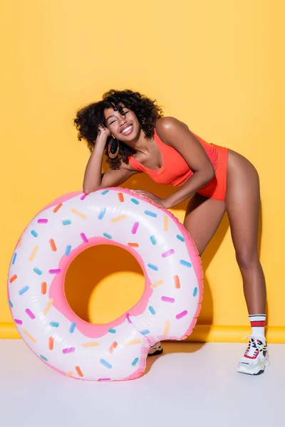 Mulher americana africana alegre em maiô sorrindo perto do anel de natação no fundo amarelo — Fotografia de Stock