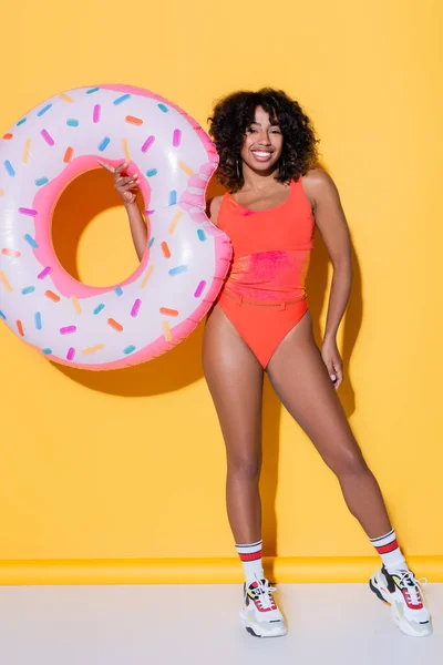 Longitud completa de la mujer afroamericana feliz en traje de baño y zapatillas de deporte con anillo de natación sobre fondo amarillo - foto de stock