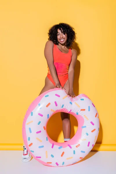 Happy african american woman in swimsuit posing near inflatable ring on yellow background — Stock Photo