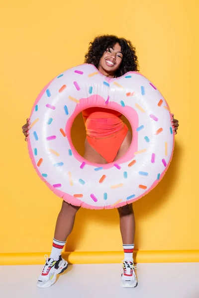 Longitud completa de la mujer afroamericana emocionada sosteniendo el anillo de natación y mirando a la cámara en el fondo amarillo - foto de stock