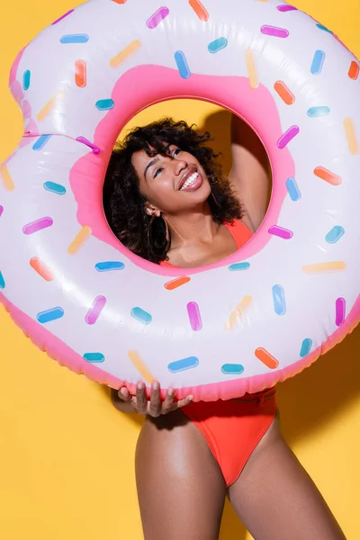 Fröhliche afrikanisch-amerikanische Frau mit lockigem Haar hält aufblasbaren Ring auf gelbem Hintergrund — Stockfoto