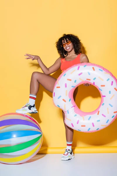 Mulher americana africana elegante posando com anel de natação e bola inflável no fundo amarelo — Fotografia de Stock