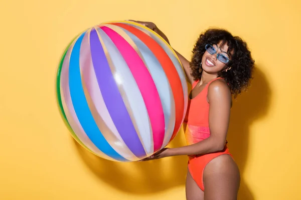 Mulher americana africana alegre em maiô e óculos de sol na moda segurando bola inflável listrada no fundo amarelo — Fotografia de Stock