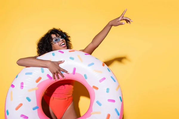 Mujer afroamericana feliz sosteniendo el anillo de natación y señalando con el dedo sobre el fondo amarillo - foto de stock