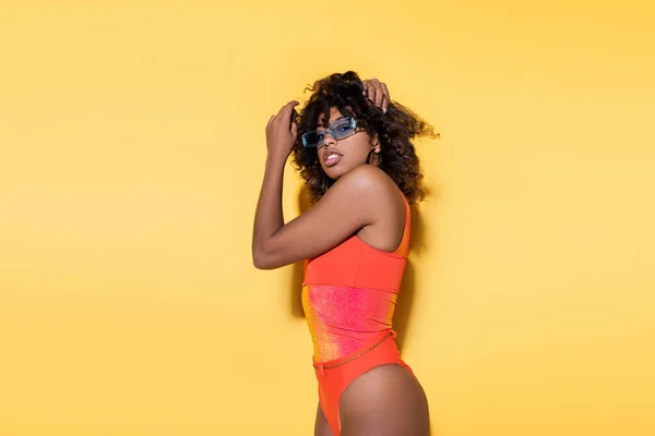 Élégante femme afro-américaine en maillot de bain rouge touchant les cheveux bouclés et regardant la caméra sur fond jaune — Photo de stock