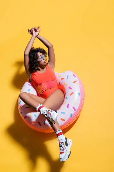 Top view of african american woman in swimsuit and stylish sunglasses sitting on swim ring on yellow background — Stock Photo