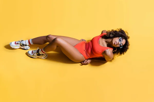 Top view of sensual african american woman in red swimsuit, sunglasses and sneakers on yellow background — Stock Photo