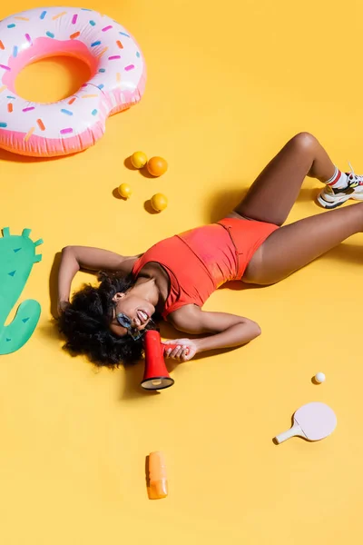 African american woman lying near fruits, swim ring and sunscreen while talking in megaphone on yellow background — Stock Photo