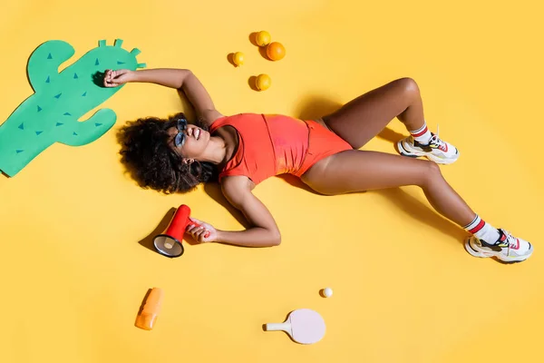 Elegante donna afroamericana sdraiata con megafono vicino a cactus taglio carta, frutta e crema solare su sfondo giallo — Foto stock