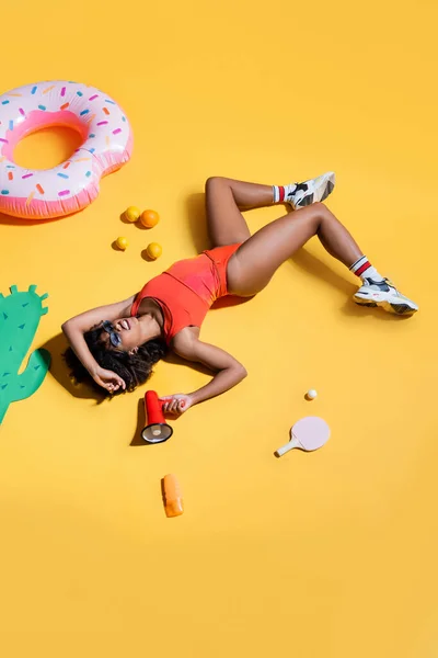 African american woman in sneakers and swimsuit lying with megaphone near fruits and swim ring on yellow background — Stock Photo