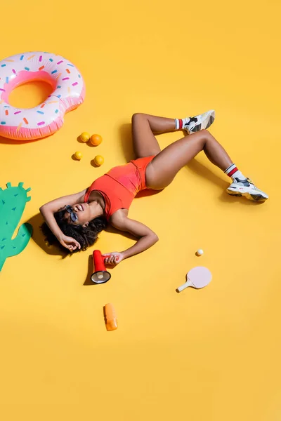 Mujer afroamericana en zapatillas y traje de baño acostado con megáfono cerca de frutas y anillo de baño sobre fondo amarillo - foto de stock