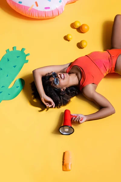 Sorridente mulher afro-americana com megafone deitado perto de papel cortar cacto e frutas no fundo amarelo — Fotografia de Stock