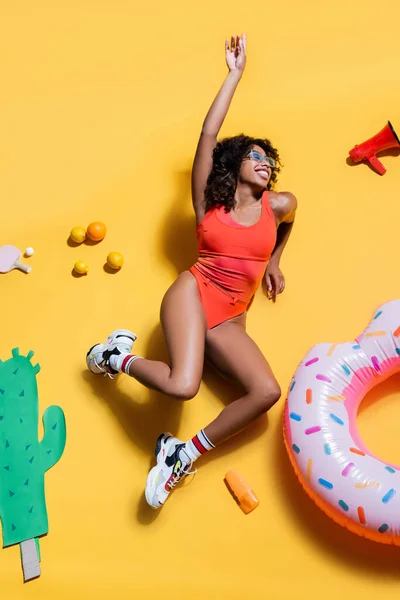 Feliz mulher afro-americana em roupa de praia perto de frutas, anel de natação e cacto corte de papel no fundo amarelo — Fotografia de Stock