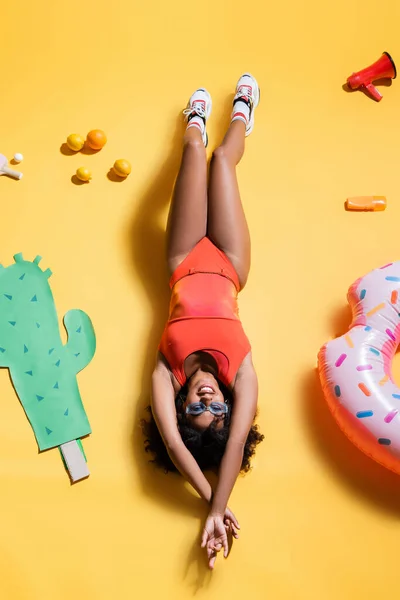 Vue du dessus de la femme afro-américaine en lunettes de soleil et maillot de bain couché près de papier coupé cactus et anneau de bain sur fond jaune — Photo de stock