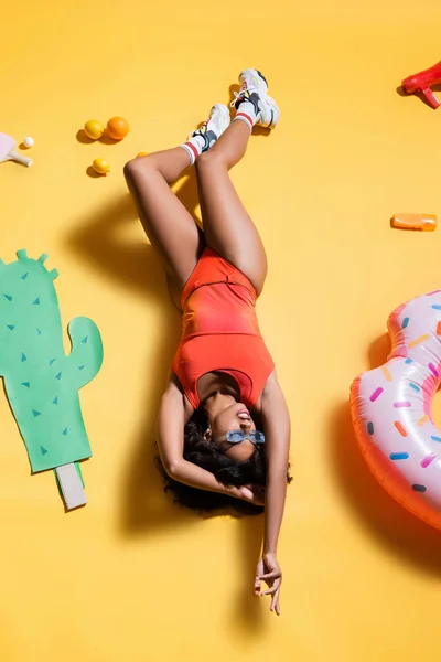 Vue du dessus de la femme afro-américaine en maillot de bain relaxant près de l'anneau de natation et du cactus coupé en papier sur fond jaune — Photo de stock