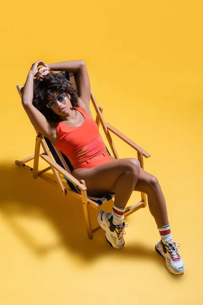 Високий кут зору афроамериканської жінки в пляжному одязі, що розслабляється в стільці на жовтому фоні — стокове фото