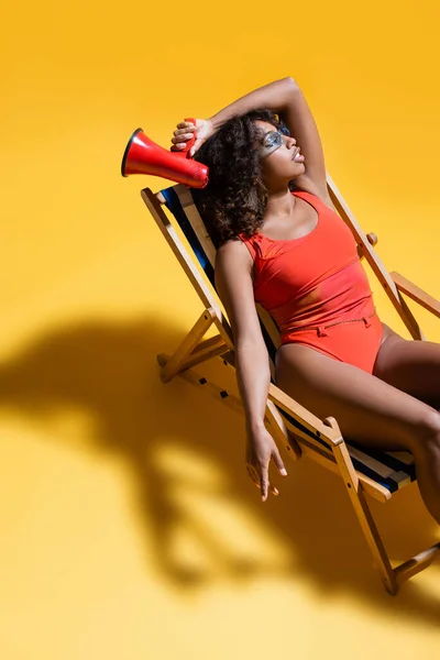 African american woman in swimsuit holding loudspeaker while sitting in deck chair on yellow background — Stock Photo