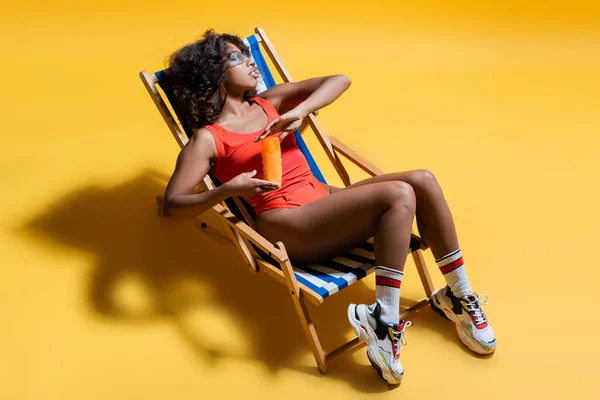 Longitud completa de la mujer afroamericana en traje de baño y zapatillas de deporte sentado en la silla de cubierta con protector solar sobre fondo amarillo - foto de stock