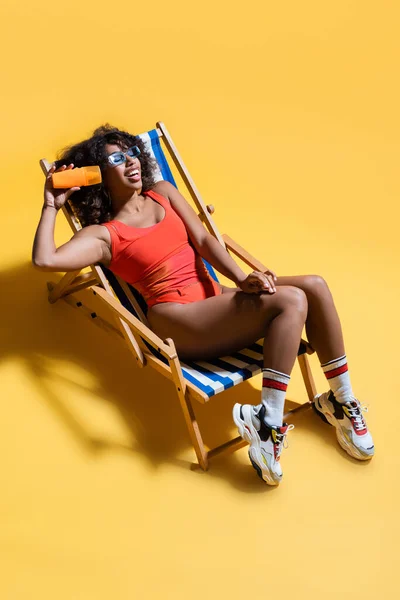 Happy african american woman in swimwear and sneakers resting in deck chair and holding sunscreen lotion on yellow — Stock Photo