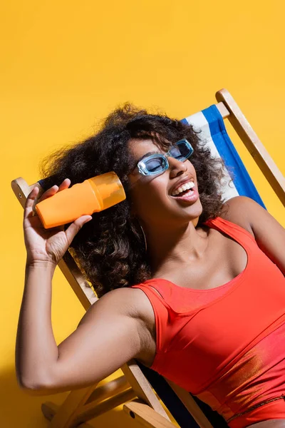 Cheerful african american woman in trendy sunglasses sitting in deck chair with sunscreen on yellow background — Stock Photo