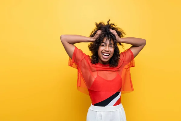 Stylish and joyful african american woman touching curly hair isolated on yellow — Stock Photo