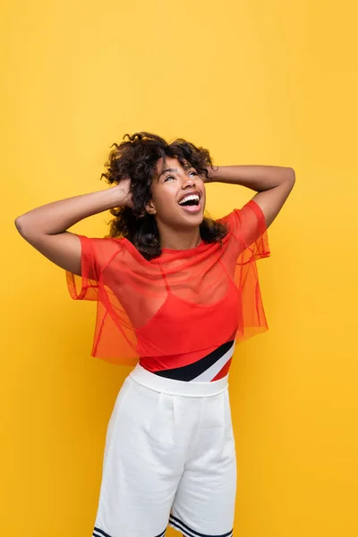 Amazed african american woman in summer outfit touching wavy hair and looking up isolated on yellow — Stock Photo