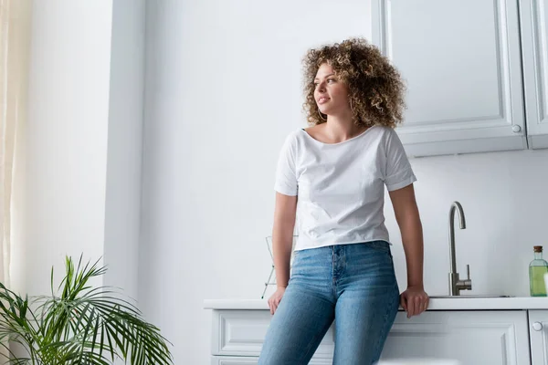 Mulher encaracolado em t-shirt branca e jeans de pé na cozinha e olhando para longe — Fotografia de Stock