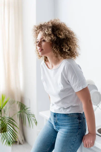 Mujer joven con el pelo rizado sonriendo mientras está de pie en la cocina — Stock Photo