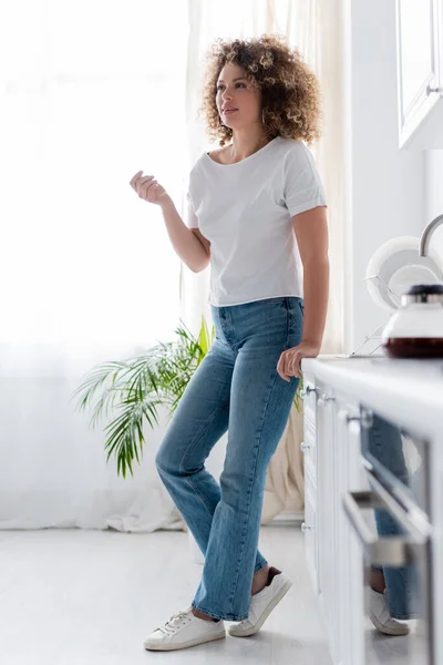 Full length view of curly woman in jeans standing in kitchen on blurred foreground — Stock Photo