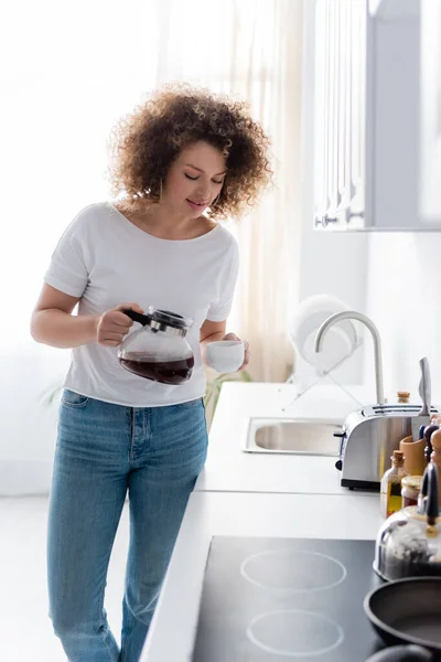 Junge lockige Frau im weißen T-Shirt, die in der Küche Kaffee einschenkt — Stockfoto