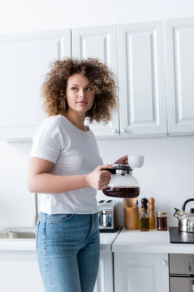 Mulher encaracolado em t-shirt branca de pé com xícara e cafeteira na cozinha — Fotografia de Stock