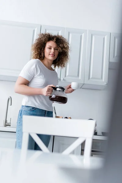 Mulher encaracolado com panela de café e xícara sorrindo para a câmera na cozinha — Fotografia de Stock